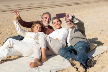 Two senior couples relaxing at seashore, lying on blanket at sandy beach and taking selfie. Bearded man holding smartphone and making shot. Modern technology, friendship, leisure concept - obrazy, fototapety, plakaty