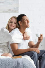 young blonde woman with closed eyes hugging happy african american boyfriend holding coffee cup in bedroom