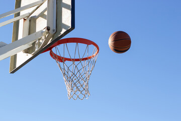 Low angle shot of basketball flying into hoop outside on sunny day. Ball thrown into basket with white backboard against blue cloudless sky. Achievement, sports concept