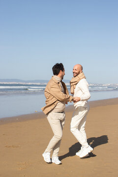 Gay Couple Spinning Around And Holding Hands On Beach. Cheerful Excited Middle Aged Men Looking At Each Other While Dancing On Sand. Dating Concept