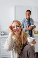 young and happy woman with closed eyes holding cup of coffee near african american boyfriend smiling on blurred background