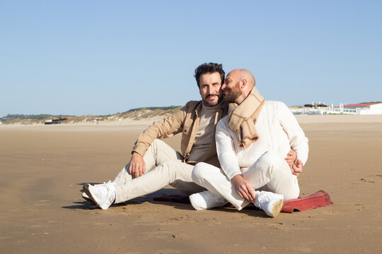 Romantic gay couple sitting on sand. Brunette bearded man hugging boyfriend on beach. LGBT tolerance concept