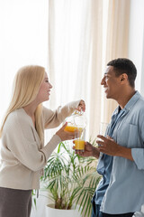 side view of blonde woman pouring orange juice near african american boyfriend holding glasses in kitchen