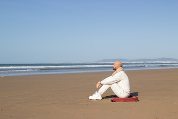 Fototapeta na wymiar Lonely middle aged man in scarf sitting on beach. Bald man in white shoes looking at ocean. Inspiration and planning concept
