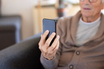 Senior man calling online via smartphone app. Closeup of aged man in cardigan sitting in armchair and using gadget at home. Technology concept