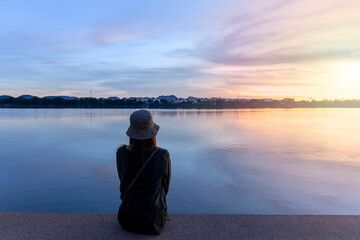 the back of a woman in a hat sitting and watching the sunrise by the river