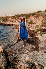 A woman in a dress, hat and with a straw bag is standing on the beach enjoying the sea. Happy summer holidays