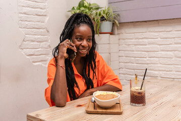 a beautiful african american woman sitting in a bar talking on the phone while enjoying acai bowl with peanut butter