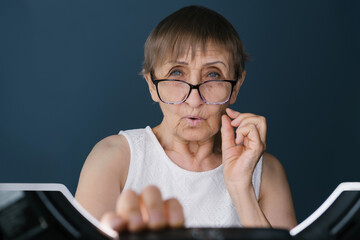 Portrait of elderly woman on dark blue background using a laptop or keyboard surprised looking at the camera over  glasses like a monitor