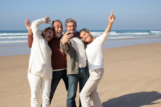 Group of senior friends having fun at the beach, standing against sea background and taking selfie. Bearded man holding smartphone and making shot. Modern technology, friendship, leisure concept