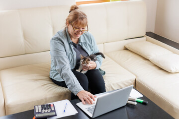 Mature woman in blue shirt sitting with a cat on her lap at the wooden table at home with laptop and notebook, working or shopping online.	
