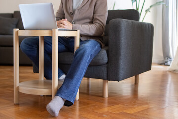 Unrecognizable man watching online course on laptop. Man in casual outfit sitting with clasped hands at coffee table in living room. E-learning concept