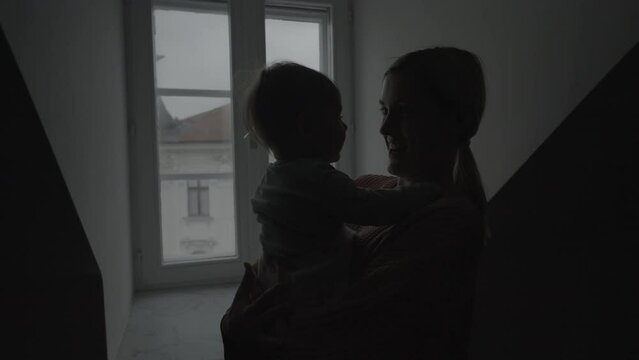Silhouette of mom holding her daughter and smiling in front of a window and looking outside on a dark and cloudy morning