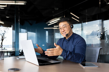 Asian with a headset is talking on video call, man inside office smiling and gesturing joyfully in...