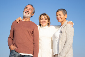 Portrait of three senior friends outdoors standing against blue sky background and smiling....