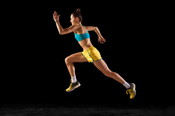 Female athlete in action. Young fitness sportive girl in sports uniform running, training isolated over dark background. Dynamic movements, running technique.