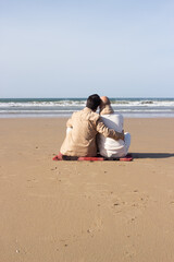 Gay couple sitting on sand outdoors. Rear view of homosexual partners embracing and looking at sea on beach. Romance concept