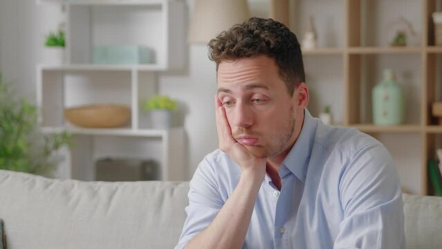 Sad bored man lean head on hand thinking seated on sofa at home alone
