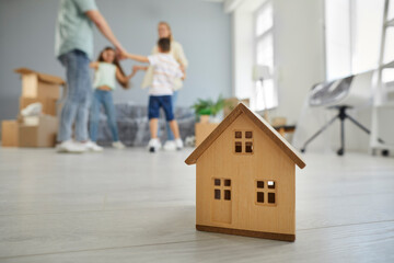 Close up shot of little wooden toy house on floor at home, with happy young family dancing and having fun in blurry background. Real estate, mortgage, buying property, insurance concept
