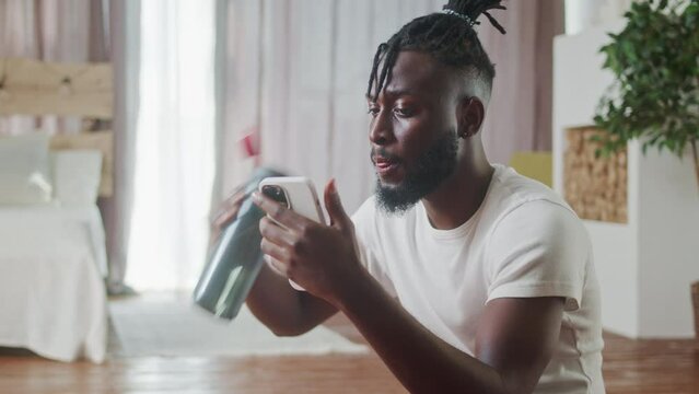 Young Beginer African American drinking bottle of water after workout, fitness, gymnastic, aerobic session. Muscullar man using phone, surfing social media, cheking news, sitiing on floor. Do sports