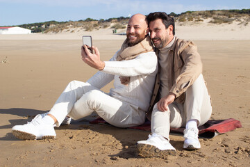 Homosexual couple taking selfie on beach. Smiling middle aged gays sitting on sand and posing together. Digital nomad and relationship concept