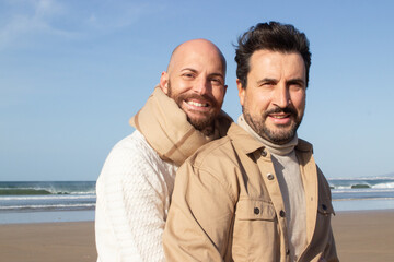 Middle aged gay hugging partner from back. Smiling bald man in scarf and his bearded boyfriend in shirt looking at camera. Homosexuality concept