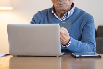 Senior man shopping online using laptop and credit card. Unrecognizable senior man sitting at table in living room, making purchases through internet, entering debit card details. E-commerce concept
