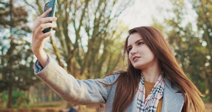 Caucasian young beautiful woman in grey coat walking in park on sunny fall day and taking selfie photo on mobile phone via webcam. Pretty cheerful girl taking selfies photos in forest among trees.