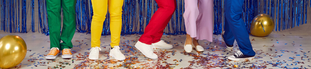 Group of five people in green, yellow, red, purple and blue trousers having fun at festive party. Banner header background. Cropped shot, legs and feet of dancing people, scattered confetti on floor