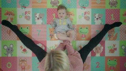 Changing diapers - Top view of a mother in a pink sweater and black leggings, sitting on a colorful mat and changing her baby daughter's nappy