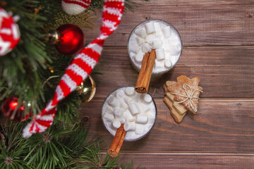 Obraz na płótnie Canvas Two christmas white drink eggnog with marshmallow and cinnamon sticks and on woodn brown background with fir-tree branches, red balls, knitted mini scarf and biscuits