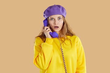 Stunned puzzled woman in purple hat and yellow sweatshirt calling phone on beige background looking at camera. Holds the purple handset of a wired phone to her face. Human sincere emotions concept.