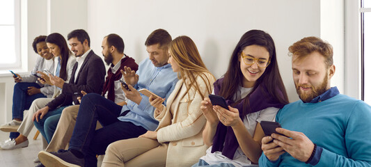Young people using smartphones. Group of different happy millennial men and women sitting in office, holding modern mobile phones, reading news, looking at memes, scrolling newsfeed and messaging