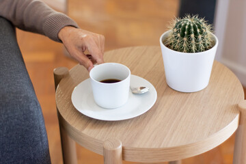 Male hand taking coffee cup from saucer. Closeup of unrecognizable man drinking espresso. Prickly cactus in pot placed on wooden table. Break concept