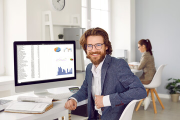 Happy male financial accountant or data analyst at work in office. Handsome bearded young man in suit and eyeglasses sitting at computer screen with graphs and charts, looking at camera and smiling - obrazy, fototapety, plakaty