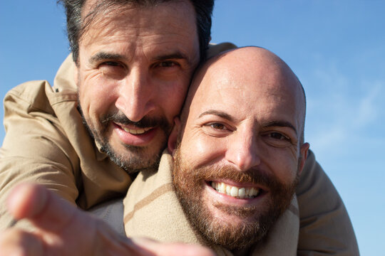 Closeup Portrait Of Happy Gay Couple Outdoors. Smiling Middle Aged Bearded Man Embracing His Boyfriend Against Blue Sky. LGBT Concept