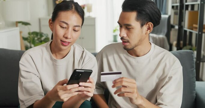 Happy young Asian couple sit on couch spend time together have fun use smartphone devices shopping use credit card online payment at home. Young husband and lifestyle domestic activity concept.
