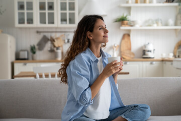 Pleased smiling woman resting on sofa with cup of tea enjoying alone time at home, looking aside...