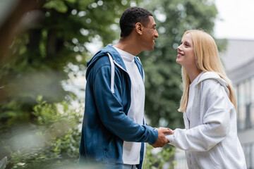 side view of blonde woman and african american man smiling at each other and holding hands outdoors