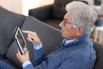 Aged man browsing internet on tablet. Serious gray-haired man in sweater sitting on sofa and using modern device. Connection concept