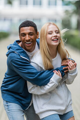 african american man laughing and hugging blonde girlfriend on blurred street in city