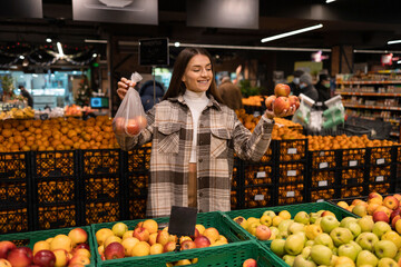 Woman in the fruit department of a grocery supermarket buys apples and thinks about whether to use a plastic bag