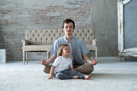 Young father trying to meditate sitting in lotus yoga pose with little son kid