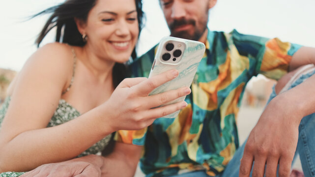 Young couple uses mobile phone while sitting on the beach on buildings background, close-up