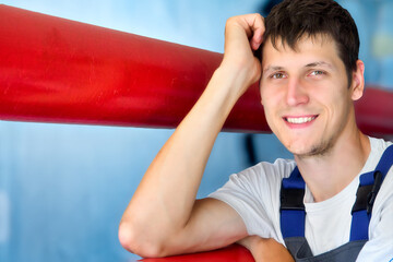 Young Caucasian worker smiles and looks into camera. Portrait of Caucasian man 30 years old. Leaning on red pipe inside room. Genuine happy worker.