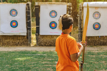 Back view of Asian boy wear face mask aims archery bow and arrow to colorful target in shooting...
