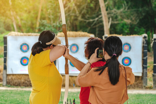 Back view of Asian woman wear visor with face mask aims archery bow and arrow to colorful target in shooting range during training and competition. Exercise and concentration with outdoor archery.