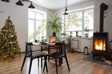 Festive interior of house is decorated for Christmas and New Year in loft style with black stove, fireplace, Christmas tree. Warm studio room with set table, burning wood, cozy and heating of home