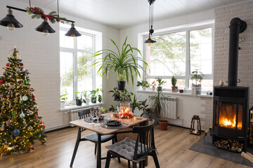 Festive interior of house is decorated for Christmas and New Year in loft style with black stove, fireplace, Christmas tree. Warm studio room with set table, burning wood, cozy and heating of home