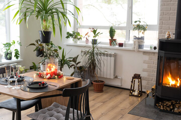 Festive interior of house is decorated for Christmas and New Year in loft style with black stove, fireplace, Christmas tree. Warm studio room with set table, burning wood, cozy and heating of home
